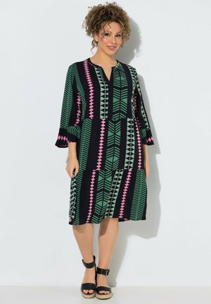 Mujer con un vestido negro hasta la rodilla con patrones geométricos verdes y rosas, de pie con sandalias negras con tiras en el tobillo frente a una pared blanca.