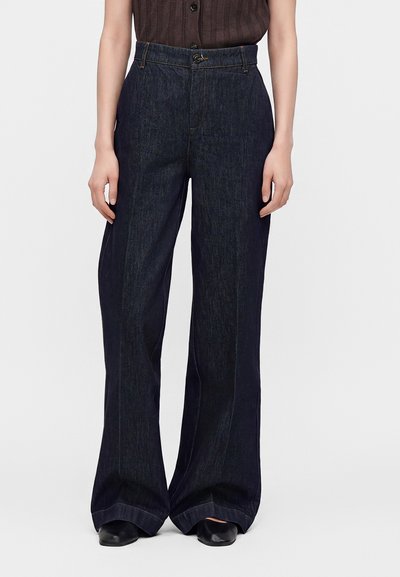 Personne portant un jean bleu foncé taille haute à jambes larges et des chaussures noires, debout devant un fond clair et uni.