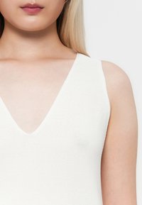 White knit tank top with a deep V-neck design, featuring thin straps and a textured fabric. Close-up view emphasizing neckline and shoulders.