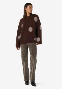 Kvinna i en brun oversized tröja med ljusgråa prickar, grå kordflottbyxor och svarta klackstövlar som står på vit bakgrund.