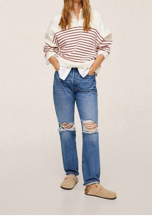 Donna che indossa un maglione crema con righe rosse, camicia bianca, jeans blu strappati e scarpe slip-on beige, in piedi con le mani nelle tasche.
