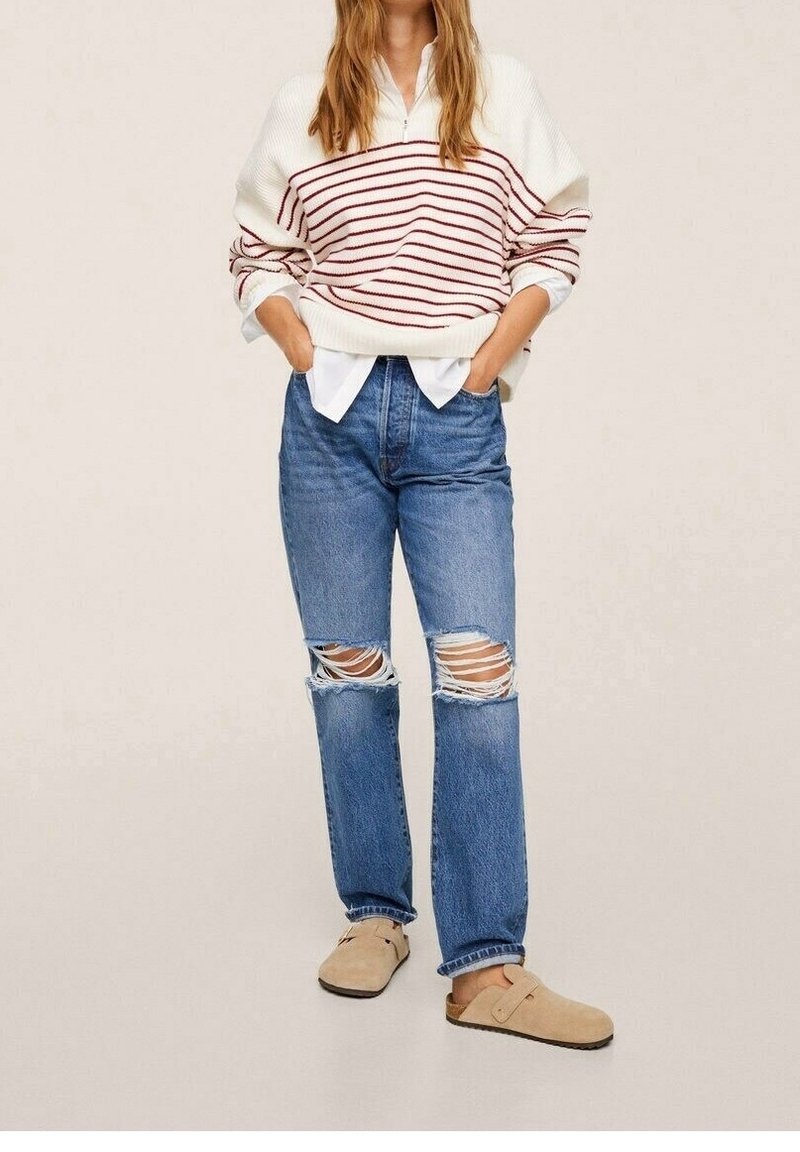 Donna che indossa un maglione crema con righe rosse, camicia bianca, jeans blu strappati e scarpe slip-on beige, in piedi con le mani nelle tasche.