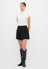 Top blanco ajustado con cuello alto, pantalones cortos negros plisados con bolsillos frontales y botas altas de cuero negro con costuras decorativas.