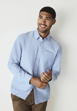 Light blue button-up shirt made of fabric with a subtle texture, featuring a chest pocket and long sleeves, paired with brown trousers.