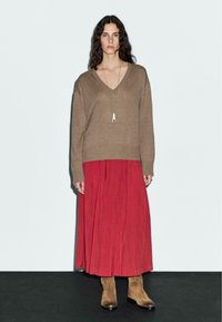 Hnědý pletený svetr s výstřihem do V, v kombinaci s plynnou červenou sukní. Nošeno s béžovými kotníkovými botami a dlouhým náhrdelníkem s přívěskem.