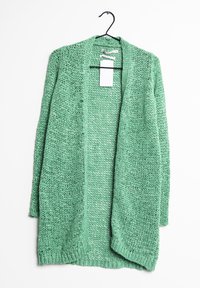 Zielony sweter robiony na drutach z otwartym przodem, lekką strukturą i luźnym wzorem splotu. Wisi na czarnym haczyku z przypiętą metką.