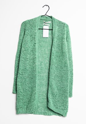 Grüne gestrickte Cardigan mit einem offenen Frontdesign, leichtem Material und lockerer Maschenstruktur. Hängt an einem schwarzen Haken mit einem angebrachten Etikett.