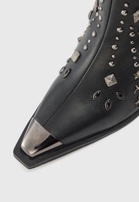 Bota de cuero negro con punta afilada, con una tapa de metal plateada y adornos decorativos de metal plateado en la parte superior.