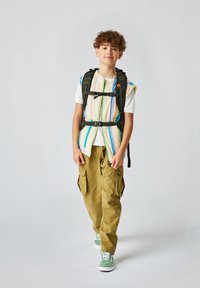 Un joven lleva una mochila negra sobre un chaleco a rayas multicolores, una camiseta blanca y pantalones cargo verdes, complementados con zapatillas verdes.