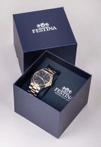 Srebrna in zlata kovinska ura s temno modrim številčnikom, prikazana v temno modri škatli. Škatla ima logotip in mehak notranji obložek.