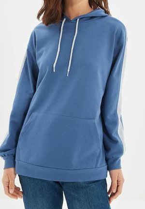 Mujer vestida con una sudadera azul con cordones blancos y rayas laterales, combinada con jeans azules, mostrada de cuello a caderas.