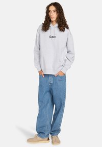 Sudadera gris clara con logotipo negro, bolsillo tipo canguro, combinada con jeans azules de corte holgado y zapatos beige. Tejido suave y diseño casual.