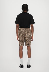 Zwarte T-shirt, achteraanzicht; tan shorts met luipaardprint, elastische tailleband, zijzakken. Model draagt witte sokken en zwarte schoenen.