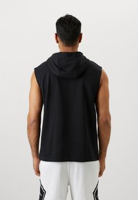 Sort ærmeløs hættetrøje med hætte, der har en glat tekstur. Bæres med hvide mesh shorts, hvilket fremhæver en afslappet atletisk stil.