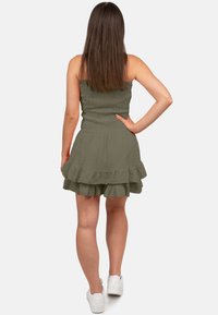 Olijfgroene strapless jurk met textuurstof, uitlopende ruches aan de onderkant en een aansluitend lijfje. Draag met witte sneakers, gezien vanaf de achterkant.