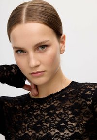 Top de encaje negro con textura transparente, mangas largas y escote redondo. El intrincado patrón presenta motivos florales y bordes festoneados.