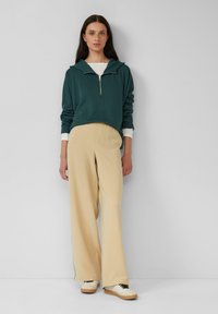 Felpa zip-up color teal con tasca frontale, pantaloni larghi beige e sneakers crema con accenti neri, stile rilassato.