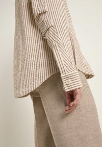 Long-sleeve shirt with textured fabric features vertical brown and white stripes, rounded hem, and buttoned cuffs. Paired with beige textured pants.
