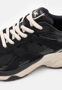 Chaussures de sport noires présentant un mélange de suède texturé et de matériaux synthétiques lisses. Soulignées par des lacets crème et une semelle amortissante.