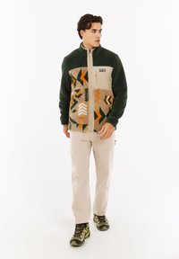Mann trägt eine gemusterte Fleecejacke mit grünen Ärmeln, beigen Hosen und Wanderschuhen, steht und schaut zur Seite vor weißem Hintergrund.