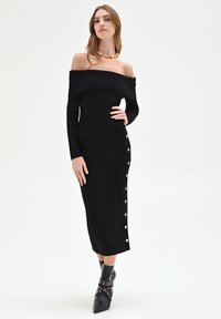 Schwarzes, geripptes, schulterfreies Kleid aus Strick mit langen Ärmeln, mittellangem Schnitt und dekorativen weißen Knöpfen entlang der Seite.