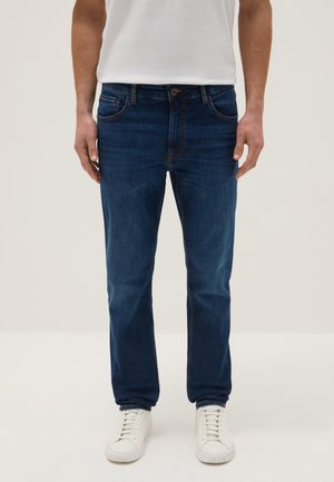 Homme portant un jean droit bleu foncé, des baskets blanches et un t-shirt blanc, debout devant un fond clair uni.