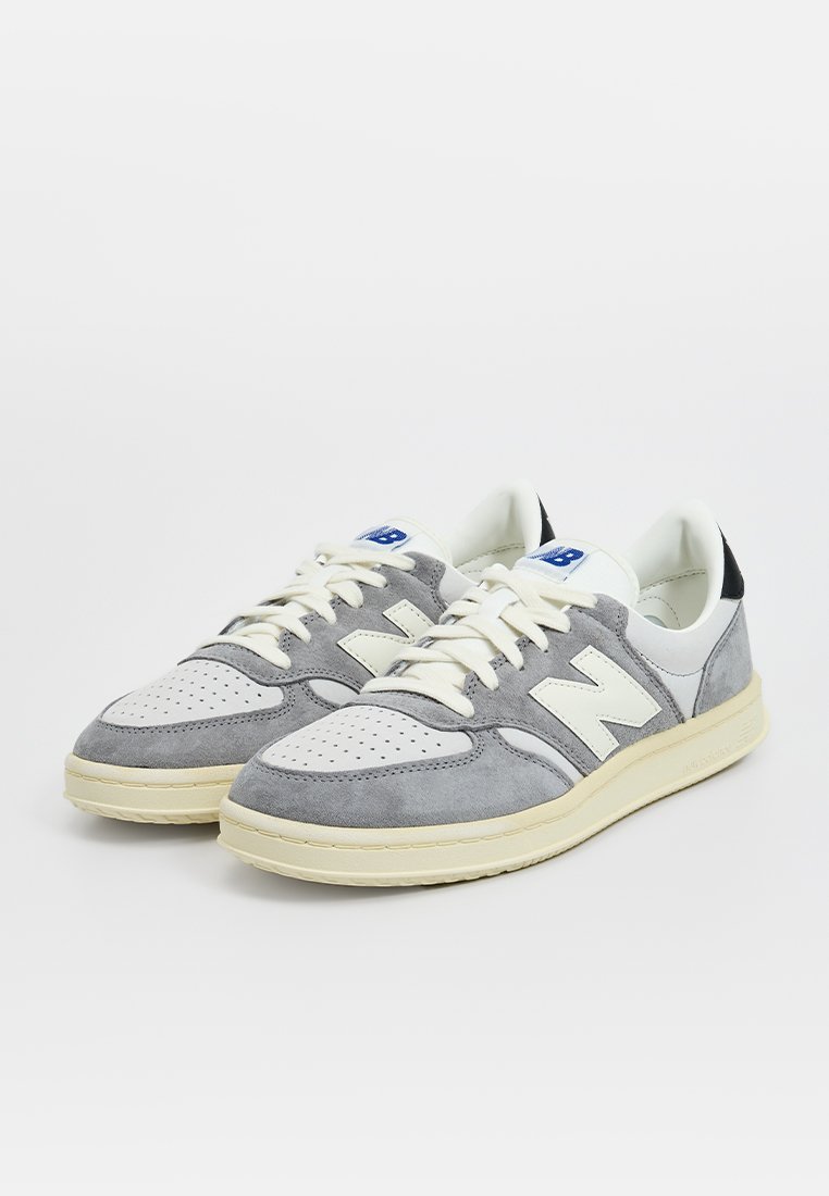 Zapatillas gris y blancas con parte superior de ante y cuero, puntera redondeada, detalles perforados y suela de goma color crema.