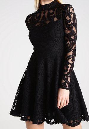 Sort blondekjole med høj hals og lange, gennemsigtige ærmer, prydet med udsmykkede blomsterdekorationer og et udvidet skørt. Teksturdetaljer i stoffet er synlige.