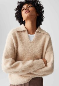 Pullover in maglina color beige chiaro, con scollo a V, colletto e una texture morbida e pelosa. Maniche lunghe con polsini a costine.