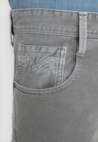 Jeans de mezclilla gris con un pequeño bolsillo frontal que presenta un diseño de costura en forma de V, bordes cortados y detalles de costura en contraste.