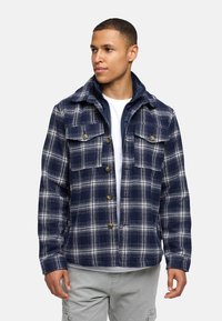 Veste en flanelle à carreaux bleu marine et blanc avec quatre poches avant, fermeture à boutons et texture douce. Superposée à un t-shirt blanc.