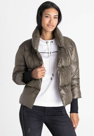 Frau trägt eine olivgrüne Steppjacke über einem weißen Hoodie und schwarze Jeans mit Nieten an den Seiten, steht vor weißem Hintergrund.