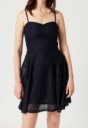 Frau trägt ein ärmelloses schwarzes Spitzenkleid mit dünnen Trägern und ausgestelltem Rock, steht vor einem einfarbigen hellen Hintergrund.
