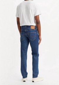 Jeans de mezclilla azul con corte recto, que incluyen bolsillos traseros y un parche de cuero marrón en la cintura. Combinados con una camiseta gris y zapatillas.