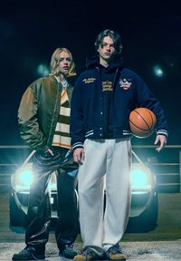 Due persone sono in piedi davanti a un'auto. Una indossa una giacca bomber verde, un maglione a righe e pantaloni neri lucidi; l'altra veste una giacca varsity blu navy, pantaloni della tuta grigi e tiene un pallone da basket.