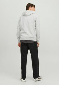 Sudadera gris con capucha y dobladillo de canalé, combinada con jeans oscuros. El modelo está de pie frente a un fondo claro, mostrando la vista posterior.