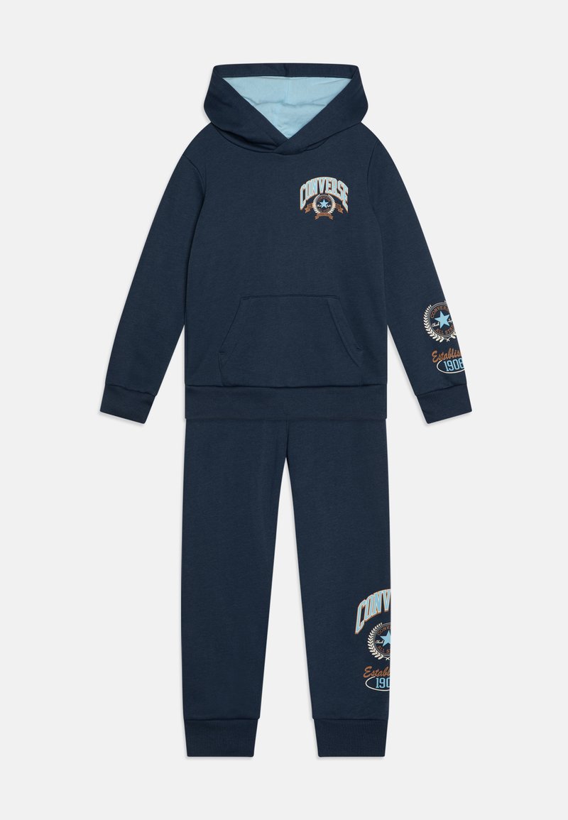 Tuta per bambini di colore blu navy di Converse, con felpa con cappuccio, tasca frontale e pantaloni coordinati, caratterizzati dai loghi del marchio sul petto, sulla manica e sulla gamba.