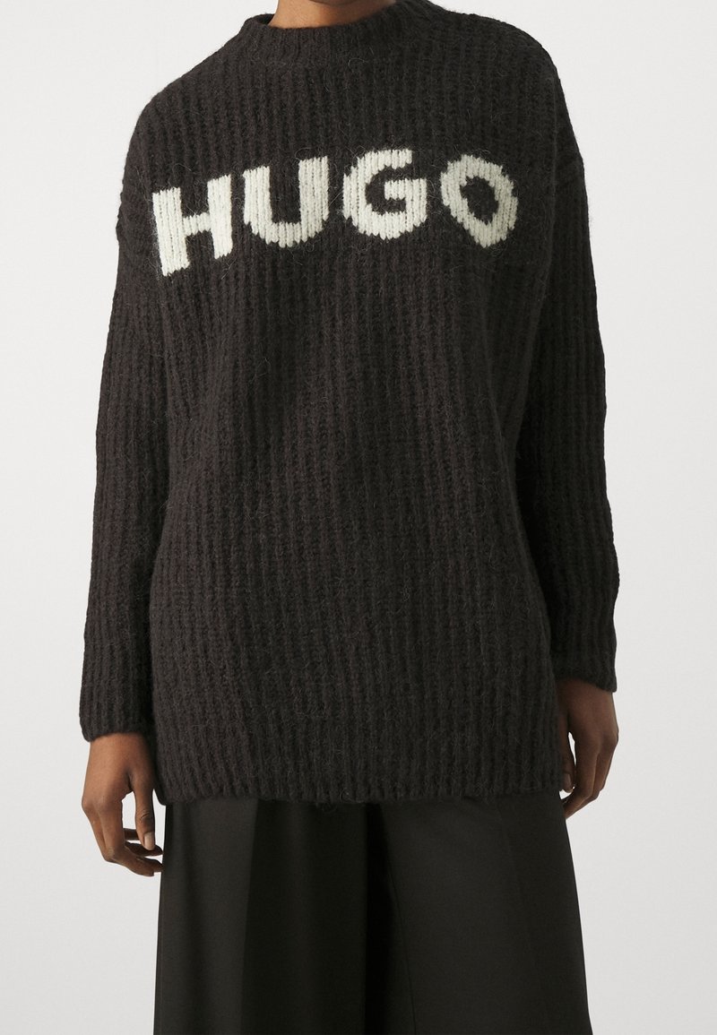 Personne portant un pull en grosse maille noire avec un texte blanc "HUGO" sur la poitrine et un pantalon noir ample sur un fond uni.