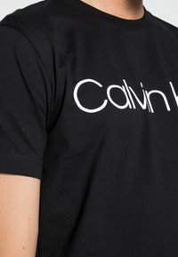 Musta puuvillapaita lyhythihaisilla, jossa edessä valkoiset tekstit "Calvin Klein". Suora pinta, standardi pyöreä kaula-aukko.