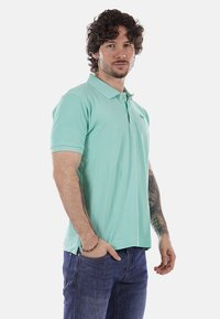Homme aux cheveux bouclés portant un polo vert menthe et un jean bleu, debout avec une main dans la poche sur un fond uni.