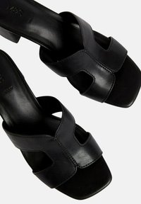 Sandalias de tacón alto de cuero negro con diseño recortado y punta cuadrada. Textura suave, costuras mínimas y forma estilizada.