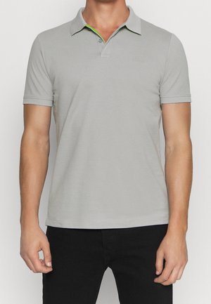 Homme portant un polo gris clair à manches courtes avec col et deux boutons, associé à un pantalon noir, debout devant un fond blanc.