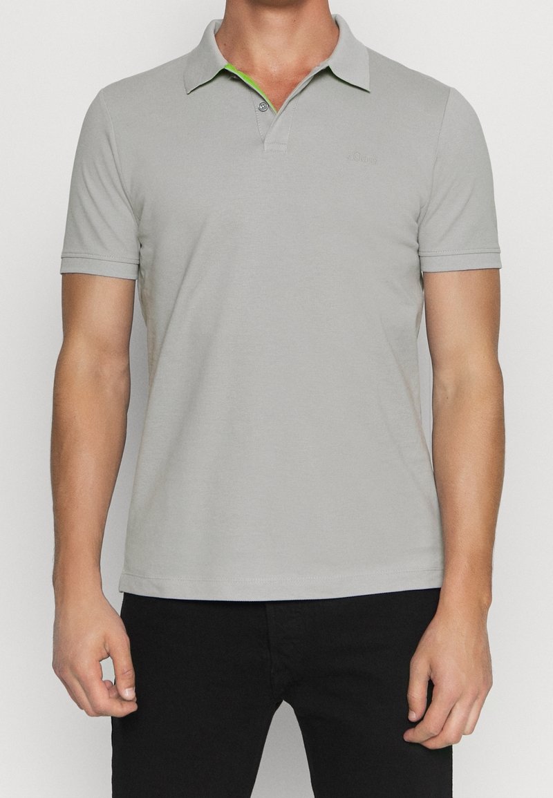 Homme portant un polo gris clair à manches courtes avec col et deux boutons, associé à un pantalon noir, debout devant un fond blanc.
