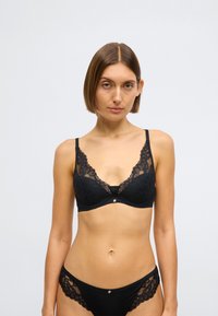 Melnā mežģīņu krūšturis ar ziedu rakstiem un regulējamām lencēm, centrā neliels metāla elements. Mīksts audums, dziļš izgriezums.