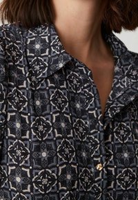 Close-up af en person iført en mørk mønstret skjorte med lyse geometriske og blomstrede designs samt en guldkneppe nær kraven.