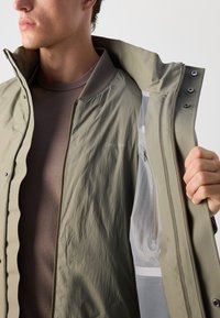 Chaqueta resistente al agua en color caqui, con cierre de cremallera, cuello de malla y bolsillos laterales. El forro interior presenta un diseño en gris claro.