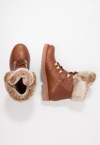 Bruna läderankelboots med en krage av fuskpäls, guldfärgad hårdvara, bruna snören och en texturerad gummisula med en kontrasterande röd rand.