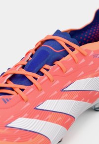 Zapato de fútbol de color rosa brillante con acentos azules, superficie texturizada, detalle de franja blanca y cordones naranjas. Presenta una forma aerodinámica para un mejor rendimiento.