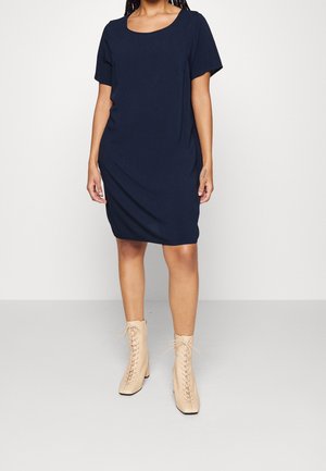 Kobieta w granatowej sukience o długości do kolan z krótkim rękawem, w beżowych, sznurowanych botkach, stojąca na tle jednolitego tła.