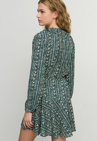 Blommönstrad klänning i teal och krämfärgad. Designen har vertikala blad, långa ärmar och en rynkad halsringning med en utställd kjol.